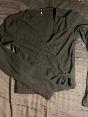 Svart omlottopp med knytband - Säljer en svart ribbad omlottopp med långa ärmar och knytband i midjan. Toppen har en smickrande v-ringning och är tillverkad i ett mjukt, stretchigt material som sitter snyggt på kroppen. Perfekt för dig som gillar stilrena och enkla plagg.