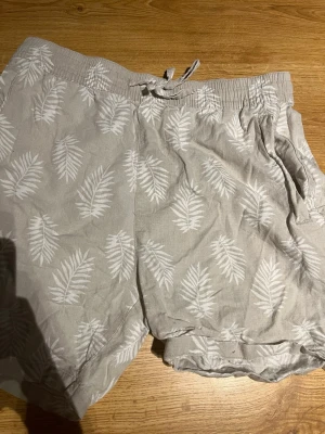 Beige shorts med palmbladsmönster - Säljer ett par beige shorts från 157 med vitt palmbladsmönster. Shortsen har elastisk midja med snörning och sidofickor. Perfekta för varma dagar och har en avslappnad passform.