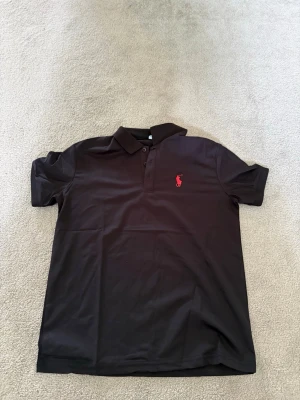 Svart pikétröja från Ralph Lauren  – Svart – Strl M – Helt ny - Klassisk svart pikétröja från Ralph Lauren med röd broderad logga på bröstet. Tröjan har krage, knappar framtill och korta ärmar. Perfekt för en stilren och enkel look. SNABB AFFÄR GER BÄTTRE PRIS 