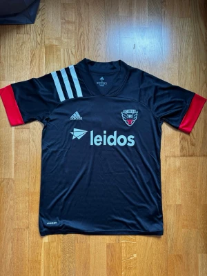 D.C. United matchtröja Adidas svart - Snygg svart D.C. United matchtröja från Adidas med röda ärmslut och ljusgrå detaljer. Tröjan har klubbmärke, sponsortryck och tre klassiska Adidas-ränder på axeln. Namnet 'Arriola' och nummer 7 på ryggen. Tillverkad i lätt och ventilerande material.