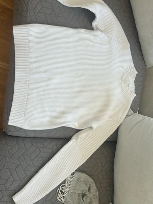 Vit ribbstickad tröja - Klassisk vit ribbstickad tröja med rund hals och långa ärmar. Tröjan har breda muddar vid ärmslut och nederkant, vilket ger en snygg och stilren look. Perfekt att matcha med jeans eller kjol för en enkel och clean outfit. Storlek S men passar M med.