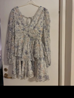 Blommig kortklänning med volang - Supersöt kortklänning med blommigt mönster i ljusa toner. Klänningen har v-ringning, knyt i ryggen, långa ärmar och volangdetaljer. Perfekt för sommarens alla tillfällen!