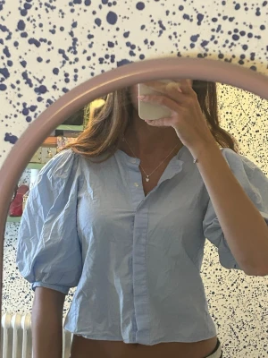 Ljusblå puffärmsblus från Zara - Trendig ljusblå blus från Zara med puffiga trekvartsärmar och knappar framtill. Blusen har en avslappnad passform och är gjord i ett tunt, vävt bomullstyg. Perfekt för en cool och stilren look.