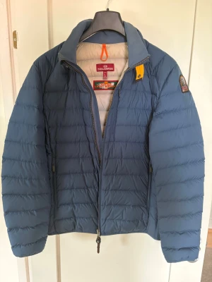 Blå dunjacka från Parajumpers - Snygg blå dunjacka från Parajumpers med quiltad design och hög krage. Jackan har dragkedja framtill, två sidofickor med dragkedja och en gul detalj vid halsen. Märkespatch på vänster ärm och lätt vadderad för extra värme. Bra skick, storlek L.