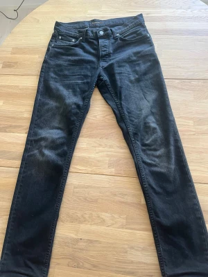 Svarta jeans från Nudie Jeans - Snygga svarta jeans från Nudie Jeans med klassisk femficksmodell och raka ben. Jeansen har en lätt tvättad look och är tillverkade i ett mjukt denimtyg som sitter skönt. Perfekta för dig som gillar stilrena och tidlösa plagg.