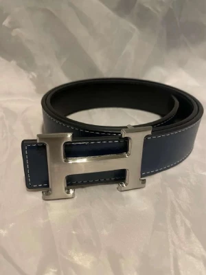 H bälte  - Snyggt blått bälte med kontrastsömmar och ett stort, silverfärgat H-format metallspänne. Bältet har en stilren design och passar perfekt till jeans eller kostymbyxor. Materialet ser ut att vara skinn.