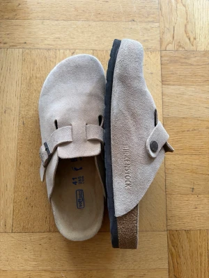 Birkenstock boston - Säljer ett par klassiska beige tofflor från Birkenstock i mjuk mocka. Modellen har en bred rem med metallspänne och präglad logga på sidan. Yttersulan är svart och ergonomiskt utformad för extra komfort. Perfekta för dig som gillar stilren och avslappnad look. Använda fåtal gånger