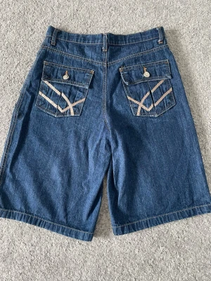Supercoola jorts! - Säljer ett par blå denimshorts med två bakfickor som har dekorativa sömmar och knappar. De har två små defekter som bara syns om man kollar väldigt nära, se bild 5