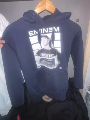 Mörkblå Eminem hoodie med tryck - Säljer en mörkblå hoodie med stor ficka framtill och huva. På bröstet finns ett svartvitt tryck med Eminem och texten slim shady  samt EMİNEM ovanför. Hoodien har långa ärmar och är gjord i mjukt material, perfekt för dig som gillar streetwear och musikstil.