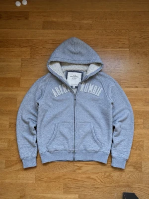 Grå hoodie med dragkedja Abercrombie & Fitch  - Mysig grå hoodie från Abercrombie & Fitch med dragkedja och stor huva. Insidan är fodrad med mjuk teddy, och framsidan har broderad text. Två fickor framtill och ribbade muddar vid ärmar och nederkant. Perfekt för chill dagar.