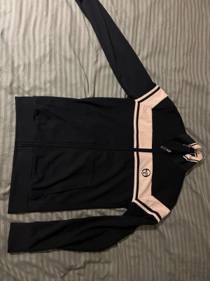Marinblå track jacket från Sergio Tacchini - Snygg marinblå track jacket från Sergio Tacchini med vita paneler och logga på bröstet. Dragkedja framtill, hög krage och två fickor. Perfekt för en sportig och retro look. Materialet är troligtvis polyester eller bomullsblandning.