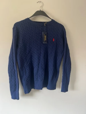 Marinblå kabelstickad tröja Polo - Snygg marinblå kabelstickad tröja från Polo Ralph Lauren med klassisk röd logga broderad på bröstet. Tröjan har rund halsringning, ribbade muddar och är gjord i ett mjukt stickat material. Perfekt för en stilren och avslappnad look. Pris går att diskuteras vid snabb affär 