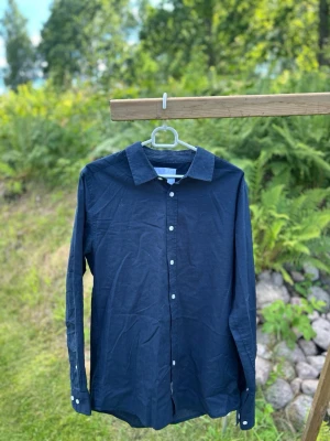 Mörkblå långärmad skjorta - Snygg mörkblå skjorta i klassisk modell med långa ärmar och vita knappar framtill. Skjortan har en rak passform och är tillverkad i ett lätt tyg, perfekt för både avslappnade och mer uppklädda tillfällen.