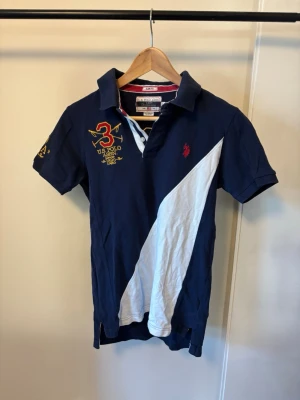 Polo Paulph Lauren Marinblå pikétröja - Snygg marinblå pikétröja med vit diagonal detalj framtill och broderade loggor i rött och gult. Klassisk krage och korta ärmar, perfekt för en sportig och avslappnad look.