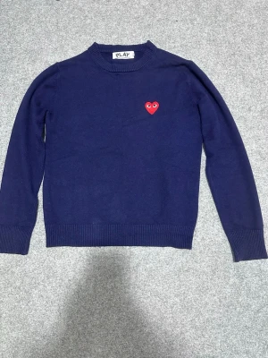 Mörkblå stickad tröja Comme des Garçons - Mörkblå stickad tröja från Comme des Garçons PLAY med det ikoniska röda hjärtat med ögon broderat på bröstet. Tröjan har rund halsringning och ribbade muddar vid ärmslut och nederkant. Perfekt för dig som gillar stilrena plagg med en twist.