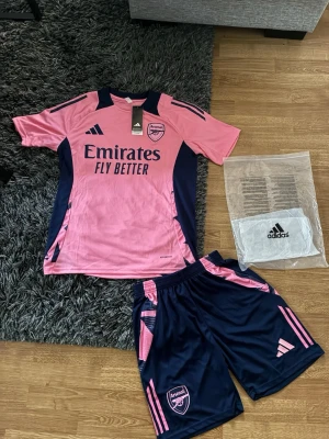 Arsenal Sett Rosa/Blå - Säljer ett Arsenal träningsset från Adidas i rosa och marinblått. Setet består av en kortärmad tröja och shorts med Arsenal-logga och Adidas-detaljer. Materialet är lätt och andas, perfekt för fotbollsträning eller gym. Snygg design med kontrastfärger och tryck.