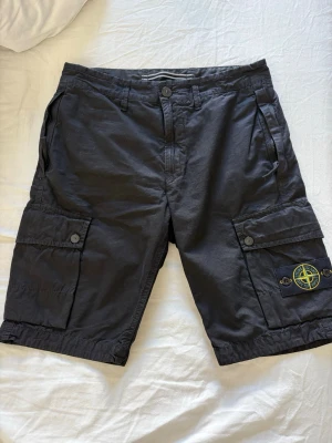 Svarta cargoshorts från Stone Island - Säljer ett par svarta cargoshorts från Stone Island med klassisk logotyp på benet. Shortsen har flera fickor med knappar. Perfekta för sommaren och har en cool streetwear-vibe. Givetvis äkta, kod finns. 