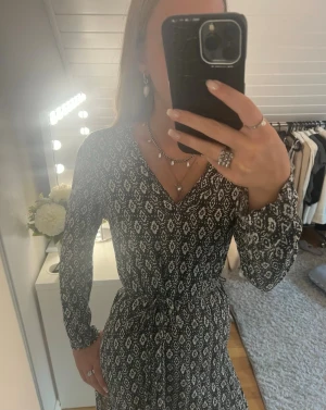 Svartvit klänning med mönster - Svartvit omlottklänning med knyt i midjan och lång ärm. Klänningen har ett diskret grafiskt mönster och v-ringad hals. Perfekt för dig som gillar en stilren och bekväm look.