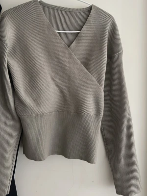 Grå ribbad omlottopp - Snygg grå ribbad omlottopp med lång ärm och v-ringad hals. Tröjan har en figurnära passform och bredare ribb nedtill som ger en modern look. Perfekt att styla med både jeans och kjol för en trendig outfit.