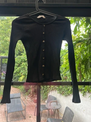 Svart ribbad långärmad från H&M - Snygg svart ribbad långärmad från H&M med knappar hela vägen framtill. Figurnära passform och rund halsringning. Perfekt att matcha med jeans eller kjol för en enkel och stilren look.