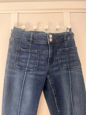 Skit snygga mörkblå bootcut jeans med fickor - Säljer ett par mörkblå bootcut jeans med raka ben och markerade sömmar framtill. Jeansen har två stora fickor fram och dubbla knappar i midjan. Materialet är klassisk denim med lätt tvättad look. Perfekta för en trendig och avslappnad stil.