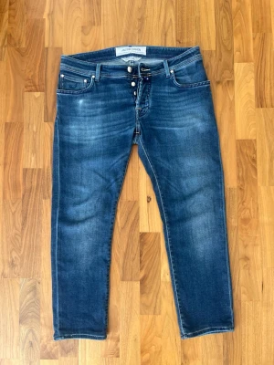 Jacob Cohen blå jeans  - Jacob Cohen jeans i modellen STYLE 622. Jeansen har en knappgylf och detaljerade sömmar. Bakfickorna har Jacob Cohens signaturdesign och en blå läderlapp med logotypen. Storlek 34 Längd 92cm Bredd 45cm