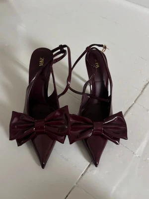 Burgundy klackskor med rosett från Zara - Säljer ett par burgundy klackskor från Zara i glansigt lackmaterial. Skorna har spetsig tå, smala remmar och en stor dekorativ rosett framtill. Klackarna är smala pch passar perfekt nu till sommaren🌸
