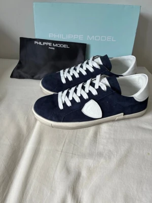 Philippe Model blåa sneakers - Mörkblå sneakers från Philippe Model i mocka med vita detaljer och vita skosnören. Klassisk låg modell med vit sula och logotyp på sidan. Perfekta för en clean och stilren look.