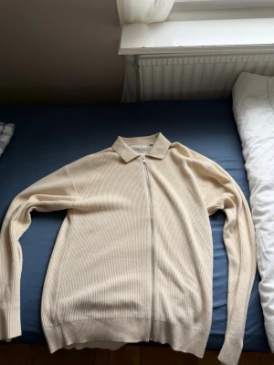 Beige ribbad ziptröja med krage - Stilren beige ziptröja i ribbat material med klassisk krage och långa ärmar. Tröjan har en hel dragkedja framtill och är tillverkad i ett mjukt, stretchigt tyg som ger en avslappnad men snygg look.