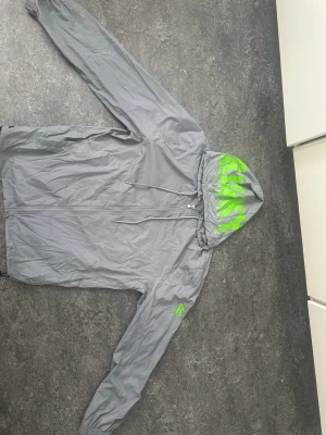 Grå vindjacka med grön detalj - Snygg grå vindjacka med huva och gröna detaljer på insidan av huvan. Jackan har dragkedja framtill, justerbar nederkant och resår vid ärmsluten. Perfekt för blåsiga dagar och enkel att packa ner i väskan.