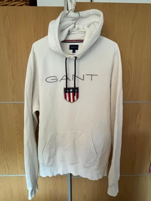 Vit hoodie från GANT med logga - Vit hoodie från GANT med stor logga och sköld på bröstet. Klassisk huva med svarta dragsnören och känguruficka framtill. Tillverkad i mjukt bomullsmaterial och har ribbade muddar vid ärmslut och nederkant. Perfekt för en avslappnad och stilren look.