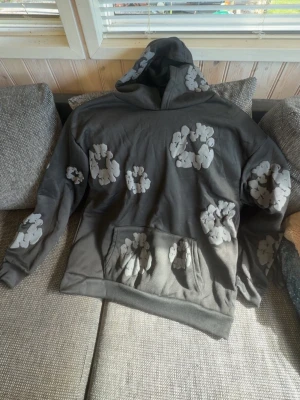 denim tears hoodie - Säljer en grå hoodie med stora och små blommönster i ljusgrått över hela tröjan. Hoodien har en klassisk huva och en stor magficka framtill. Perfekt för dig som vill ha en unik och bekväm stil. Materialet känns mjukt och skönt.