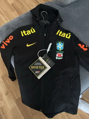 Svart Brasil Nike vindjacka Gore-Tex - Priset kan diskuteras. Svart vindjacka från Nike med Brasilien landslagslogga, gula och orange tryck samt sponsormärken. Jackan har huva, dragkedja och är tillverkad i vattentätt Gore-Tex-material. Perfekt för dig som gillar sportig stil och vill hålla dig torr.