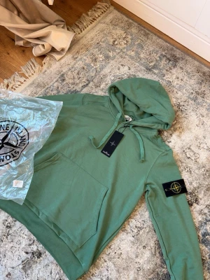 Grön hoodie från Stone Island - Snygg grön hoodie från Stone Island med klassisk patch på ärmen och stor känguruficka framtill. Tröjan har huva med dragsko och är gjord i mjuk bomull. Perfekt för dig som vill ha en stilren och bekväm look.