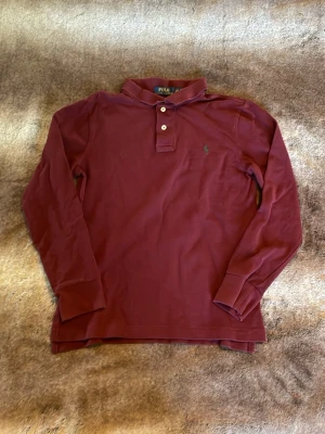 Vinröd långärmad piké från Polo Ralph Lauren - Snygg vinröd långärmad pikétröja från Polo Ralph Lauren med klassisk krage och knappar framtill. Tröjan har en liten broderad logga på bröstet och är tillverkad i mjuk bomull. Perfekt för en avslappnad men stilren look.
