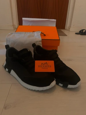 Svarta Hermes sneaker - Snygga svarta sneakers från Hermès med chunky vit sula och svarta skosnören. Skorna har en modern design med detaljer i mocka och textil, samt en ikonisk orange Hermès-box och dustbag medföljer. Perfekta för dig som vill ha något exklusivt och trendigt. Vänstra skon har en liten skada där baka men inget som syns eller påverkar skon på ett stort sätt. Hör av er om ni har några frågor🤗 