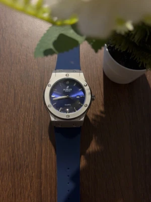 Klocka  - Stilren klocka med blått armband och matchande blå urtavla. Boetten är silverfärgad med synliga skruvar och tydliga markeringar. Klockan har datumvisning och klassisk rund form, perfekt för dig som gillar en modern accessoar.