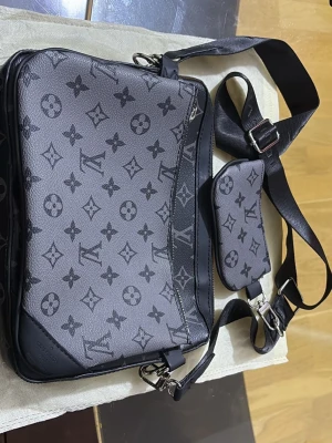 Svart axelväska med monogram  - Snygg svart axelväska med klassiskt monogrammönster och justerbar axelrem. Väskan har en extra liten ficka som sitter fast på remmen och dragkedja upptill. Tillverkad i konstläder med detaljer i svart. Perfekt för dig som vill ha en trendig och praktisk väska.