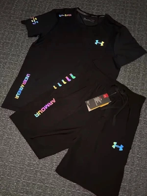 Svart träningsset från Under Armour - Säljer ett par svarta shorts från Under Armour med färgglada loggor och detaljer. Shortsen har snörning i midjan och är tillverkade i ett mjukt och lätt material som passar perfekt för träning eller chill. Stilen är sportig och modern med regnbågsfärgade tryck. Storlek S, helt ny