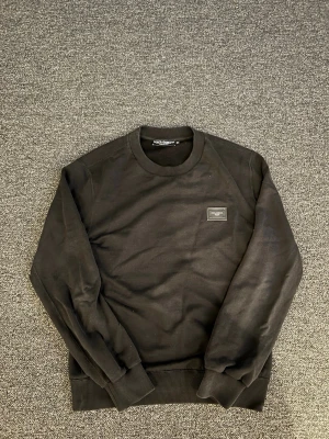 Svart sweatshirt från Dolce & Gabbana - Stilren svart sweatshirt från Dolce & Gabbana med rund halsringning och diskret logopatch på bröstet. Tröjan har ribbade muddar vid ärmslut och nederkant, perfekt för en clean och klassisk look.
