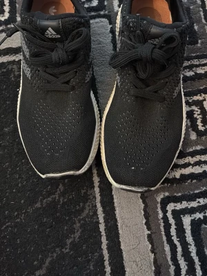 Svarta Adidas sneakers med vit sula - Snygga svarta Adidas sneakers med stickad ovandel och vita sulor. Skorna har klassisk snörning och diskreta grå detaljer på sidorna. Adidas-logga syns på plösen och innersulan. Perfekta för en sportig och avslappnad stil.