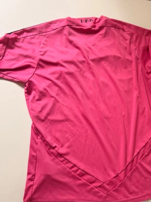 Rosa Adidas fotbollströja ifk - Säljer en rosa fotbollströja från Adidas med svarta detaljer och tryck. Tröjan har svarta ränder på axlarna, klubbmärke på bröstet och stor ICA-logga framtill. Tillverkad i funktionsmaterial (ClimaLite) som andas, perfekt för träning eller match.