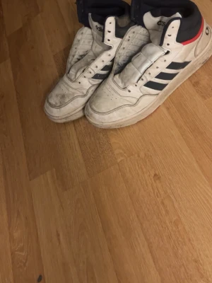 Vita Adidas sneakers med svarta ränder - Säljer ett par klassiska vita Adidas sneakers med svarta ränder och röda detaljer vid hälen. Skorna har hög modell och är tillverkade i läder med textilfoder. Perfekta för dig som gillar retrostil och streetwear.