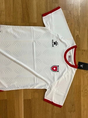 Vit Jordan matchtröja från Kelme - Snygg vit matchtröja från Kelme med Jordaniens landslagsemblem på bröstet. Tröjan har röda detaljer vid krage och ärmslut samt diskret mönster i tyget. Tillverkad i lätt och ventilerande material, perfekt för träning eller match.             Fraktar samma eller dagen efter du köpt!!! Endast en kvar i lager!!!