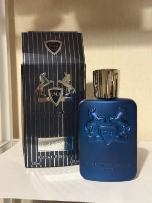 Parfums de Marly Layton 125ml - Lyxig parfym i en mattblå flaska med silverfärgad kork och upphöjda hästmotiv. Flaskan rymmer 125 ml och kommer i en elegant randig kartong med silverdetaljer. Doften är en del av Royal Essence-kollektionen och utstrålar exklusivitet.