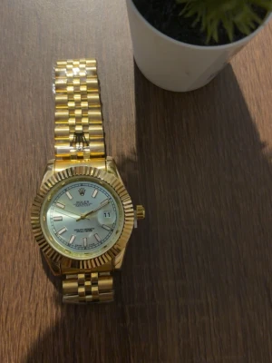 Klocka  - Stilren guldfärgad klocka med klassisk länk och räfflad bezel. Urtavlan är ljus med tydliga index och datumvisning. Perfekt accessoar för dig som vill sticka ut med en lyxig och tidlös look.