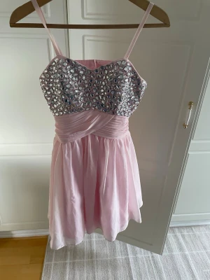 Rosa kort klänning med paljetter - Söt ljusrosa kort klänning med smala axelband och glittrig paljettdekor upptill. Livet har omlott-detalj och kjolen är lätt utsvängd i ett tunt, svepande material. Perfekt för speciella tillfällen när du vill sticka ut lite extra.