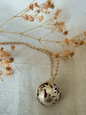 Vintage Skultuna halsband  - Elegant vintage halsband från Skultuna med dekorativt silverhänge som omsluter en bärnstensfärgad vikingpärla. Den öppna designen ger ett vackert ljusspel och framhäver pärlan på ett stilfullt sätt. Halsbandet är i fint vintage-skick med bruksslitage. Se bilder för detaljer och skick. Ett vackert smycke för både samlare och den som uppskattar klassisk skandinavisk design med historisk inspiration. Halskedjan är mest sannolikt ej original Skultuna utan senare guldplaterad ersättningskedja. 