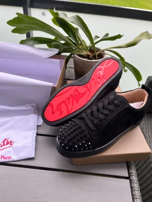 Christian Louboutin med spikar svart - Säljer ett par svarta sneakers från Christian Louboutin i mocka med svarta nitar på tån och den ikoniska röda sulan. Skorna har svarta skosnören och en låg profil som ger en clean och edgy look.