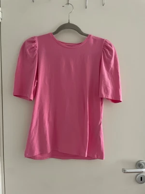 Rosa topp med puffärm - Snygg rosa topp med puffiga ärmar och rund halsringning. Tillverkad i mjuk bomull som känns skön mot huden. Perfekt för dig som vill ha en enkel men trendig look med lite extra volym på axlarna.